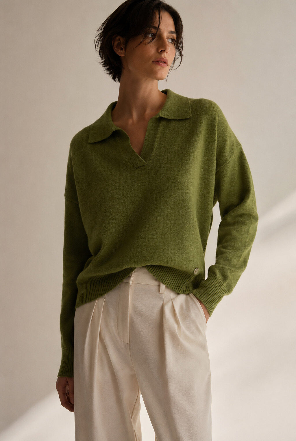 A woman wearing an olive green knit polo sweater and cream-colored trousers, standing against a warm beige background with soft shadows.