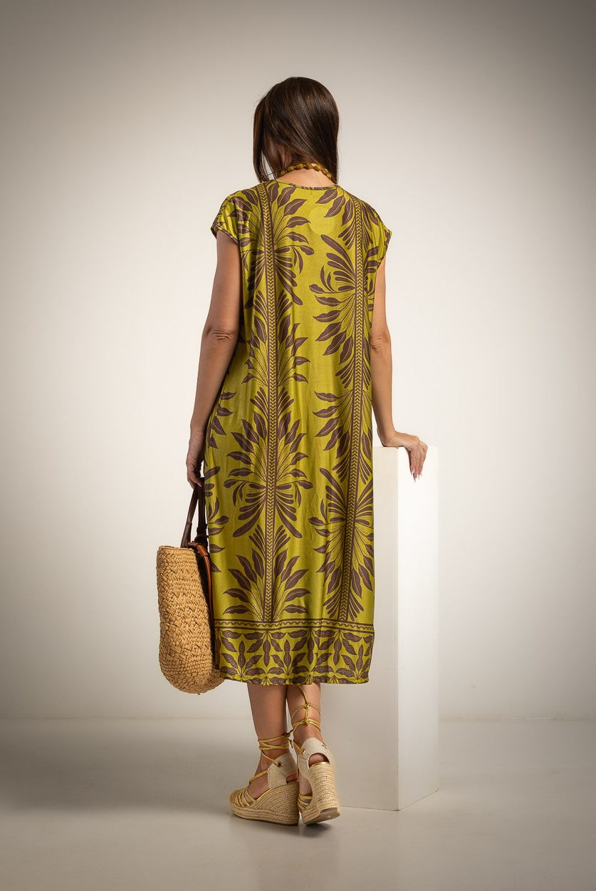 A woman stands with her back to the camera wearing a sleeveless olive green midi dress with a brown palm leaf pattern, holding a woven straw tote bag, set against a neutral studio background.