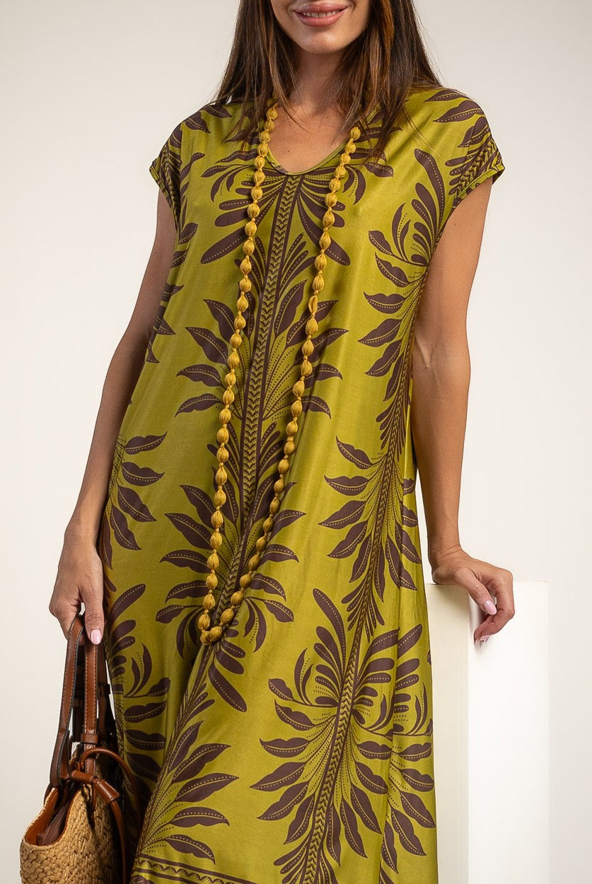 A woman wearing a short-sleeve kaftan dress in olive green with a dark brown botanical leaf pattern, accessorized with a long beaded necklace and holding a woven handbag.