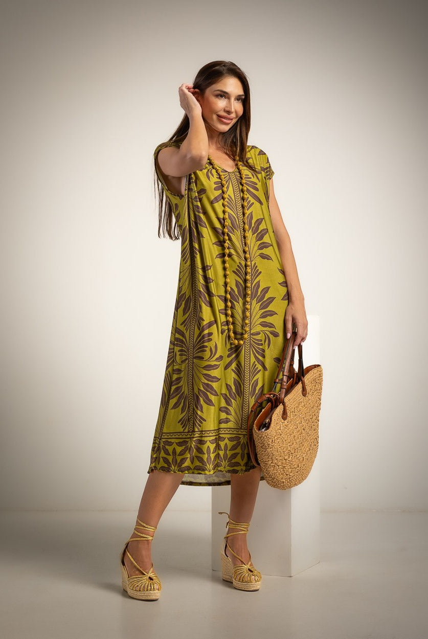 A woman models a sleeveless olive green midi dress with a brown botanical pattern, holding a woven straw tote bag and standing on a white pedestal against a neutral studio background.