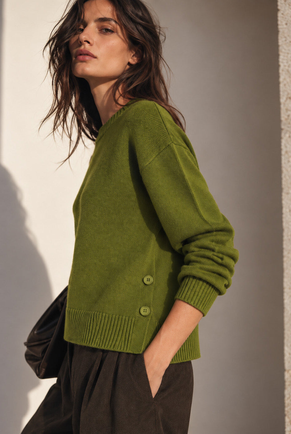 A woman wearing an olive green knit sweater with buttons on the side, paired with dark brown trousers, standing against a textured wall with warm sunlight and shadows.