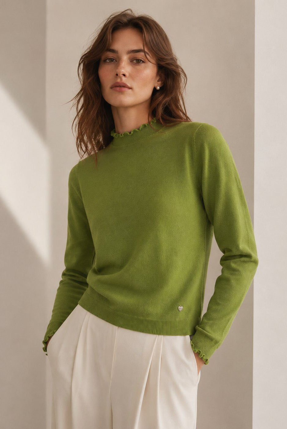 A woman wearing a light olive green knit sweater with a scalloped mock neck and white trousers, standing in a minimalist beige room with soft diagonal shadows.