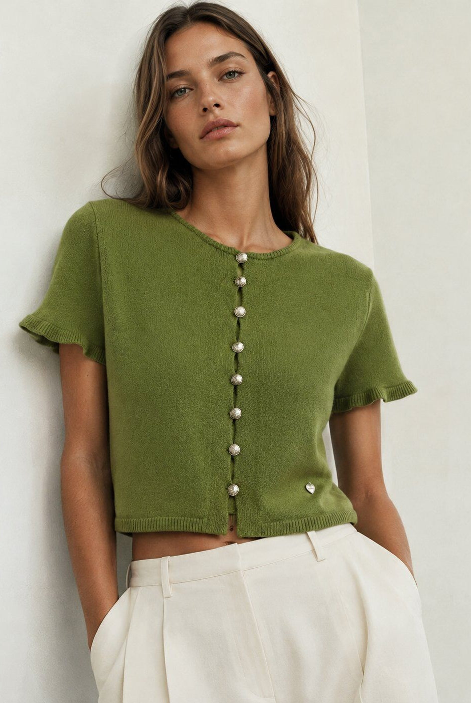A woman wearing a cropped olive green knit top with short sleeves and a vertical row of pearl-like buttons down the front, paired with white trousers, standing against a plain off-white wall.