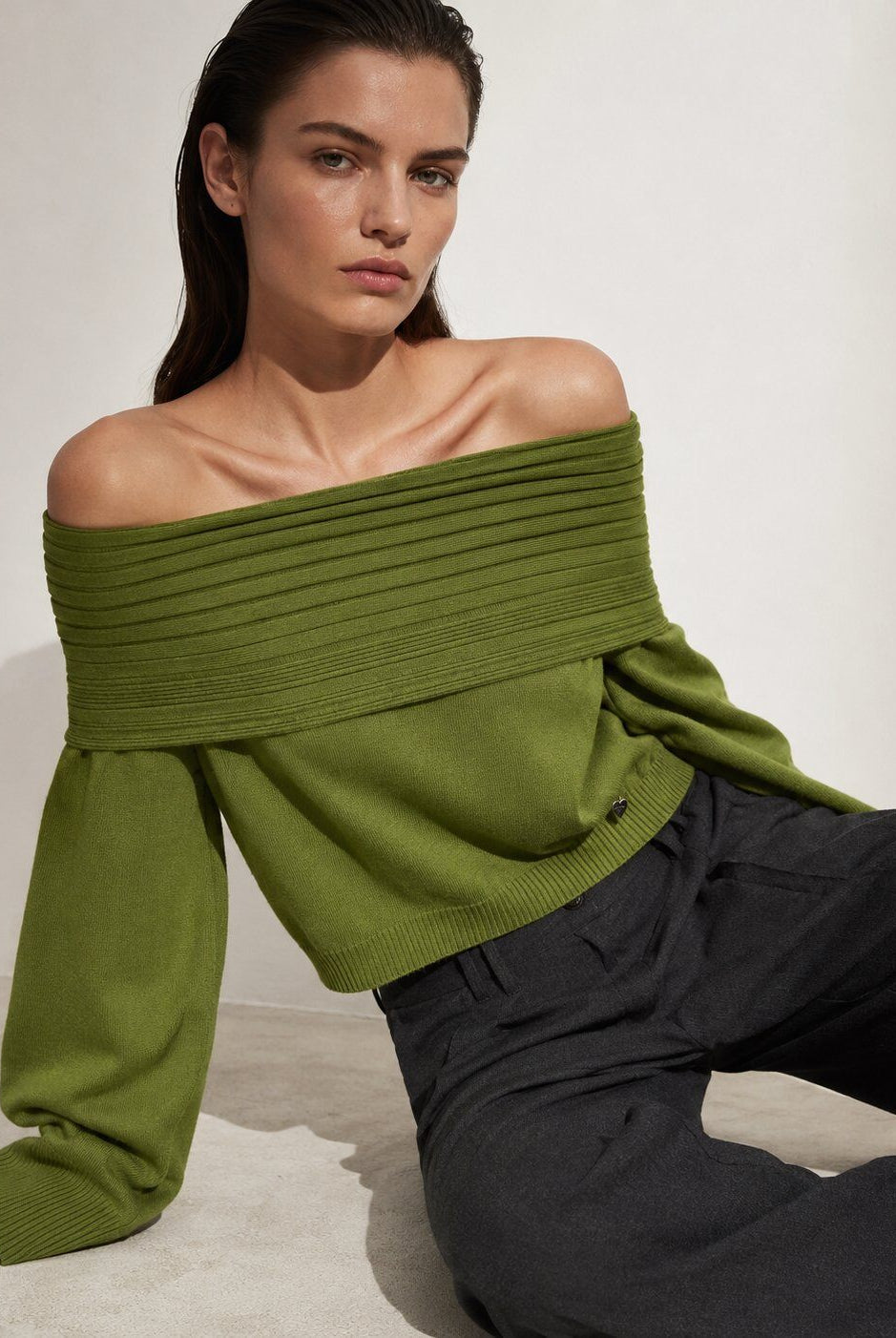A woman wearing an olive green off-the-shoulder ribbed knit sweater and dark charcoal trousers, posing against a neutral off-white background.