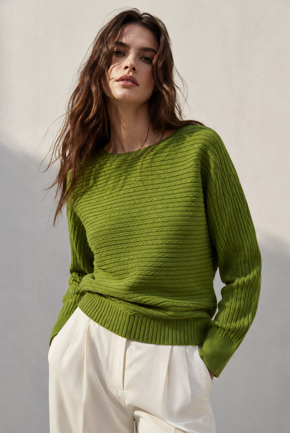 A woman wearing a textured olive green knit sweater and white trousers, standing against a neutral light grey wall.