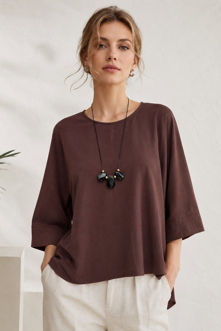 A woman wearing a loose-fitting, dark brown short-sleeve top with three-quarter sleeves, paired with off-white trousers and a black pendant necklace