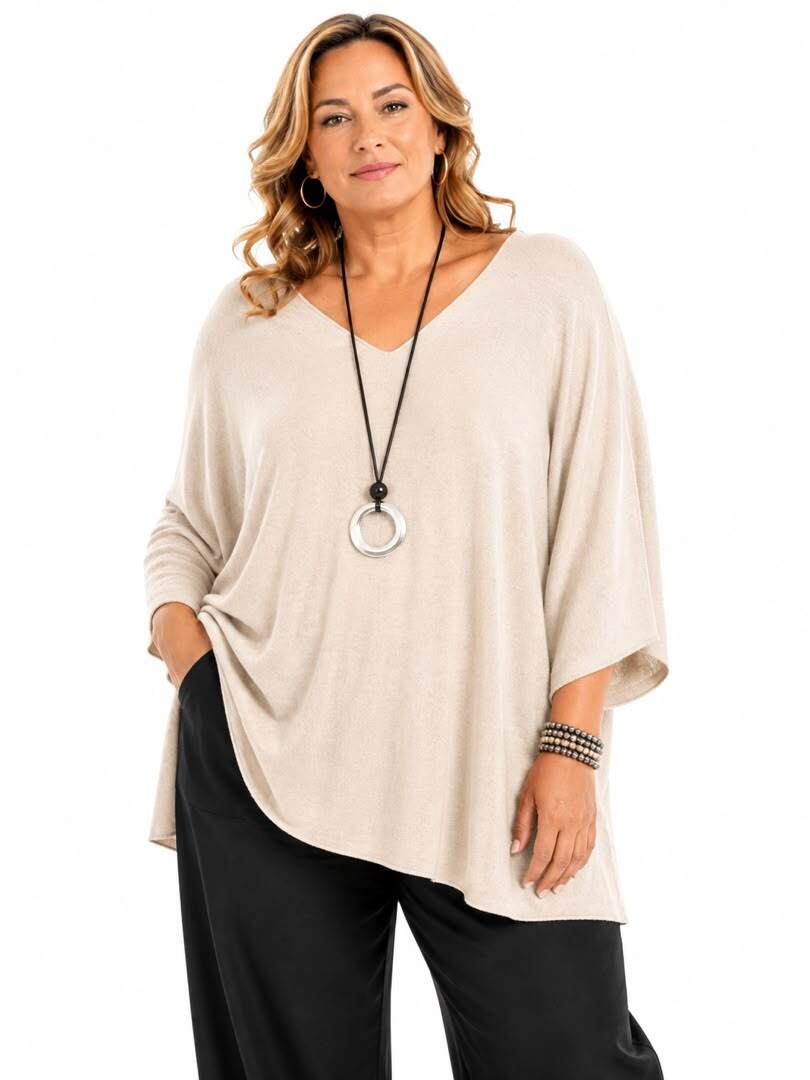 A woman modeling a light beige, loose-fitting V-neck top with three-quarter sleeves, paired with black pants and a long silver pendant necklace, against a plain white background.