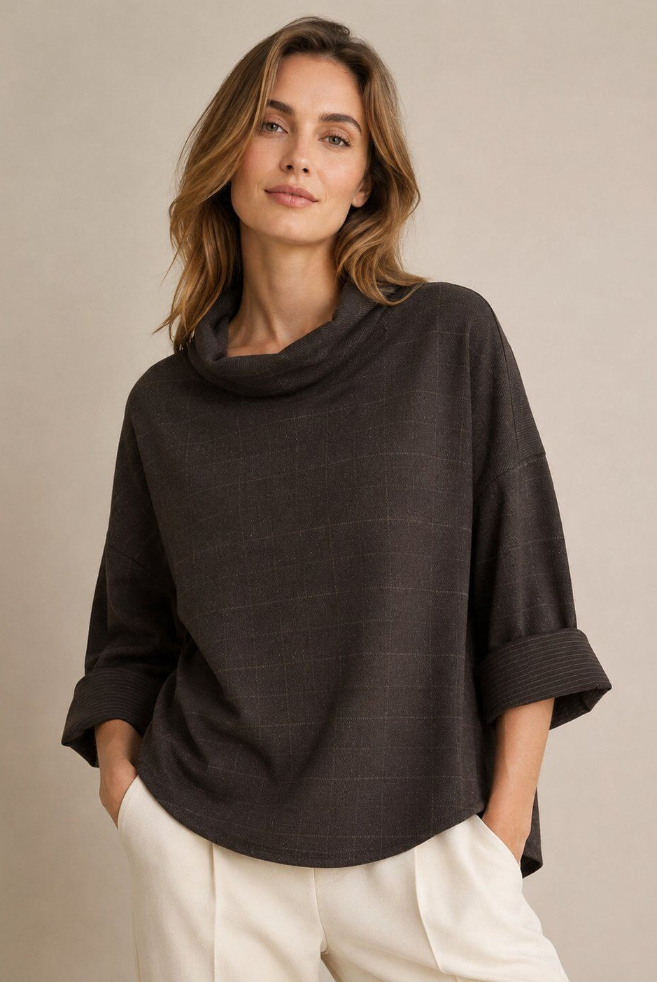 A woman wearing a dark brown cowl-neck poncho top with rolled three-quarter sleeves and white trousers, standing against a neutral beige studio background.