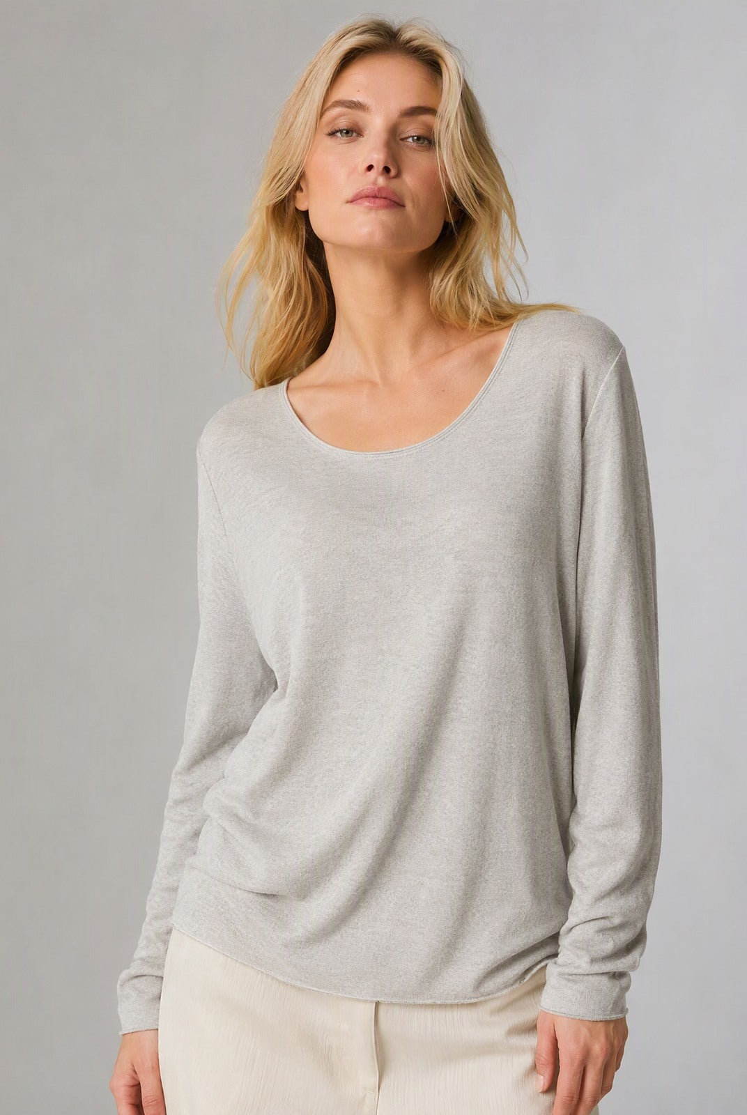 A woman modeling a light beige long-sleeve scoop-neck knit top, paired with cream pants, against a solid light gray studio background.