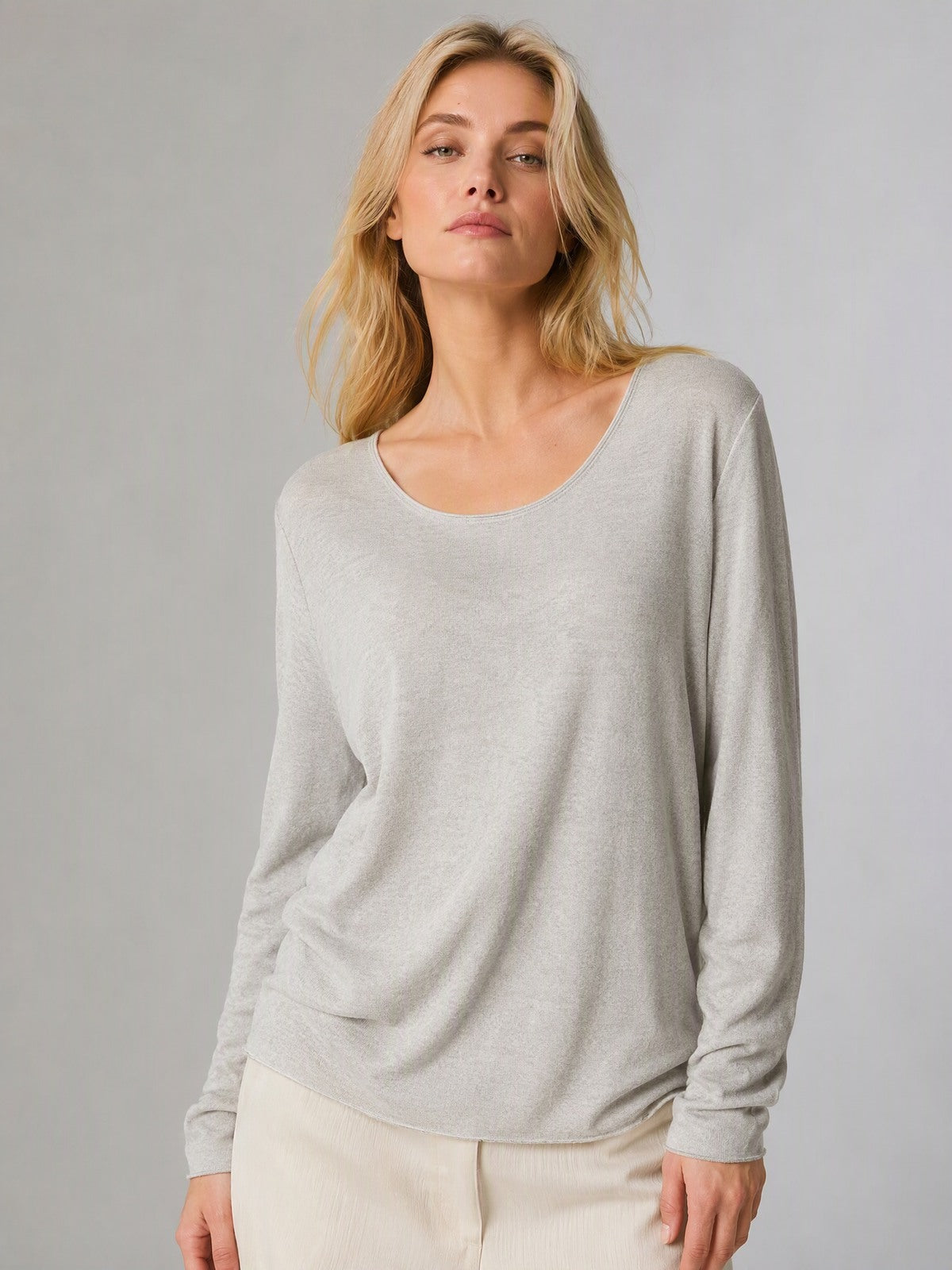 A woman modeling a light beige long-sleeve scoop-neck knit top, paired with cream pants, against a solid light gray studio background.