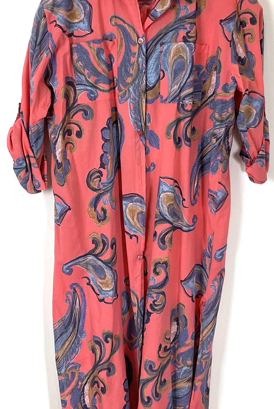 A coral pink long-sleeve button-up shirt dress with a blue, purple, and gold paisley pattern, shown hanging against a plain white background.
