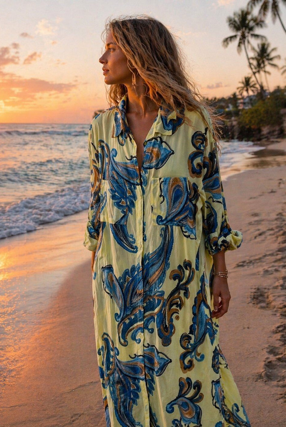 A woman stands on a beach at sunset wearing a long-sleeve button-front dress with a pale yellow base and a bold blue and dark brown ornamental print.