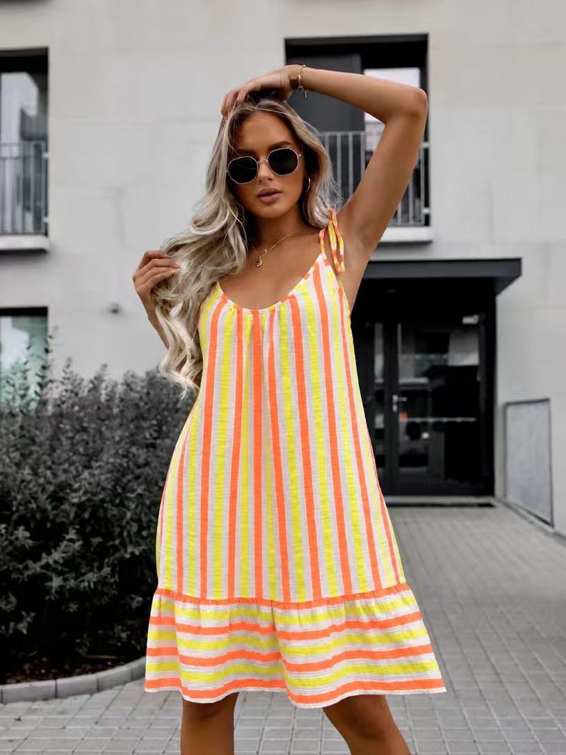 A woman wearing a sleeveless midi dress with vertical neon yellow, orange, and white stripes, styled with round sunglasses, posing outdoors in front of a modern building.