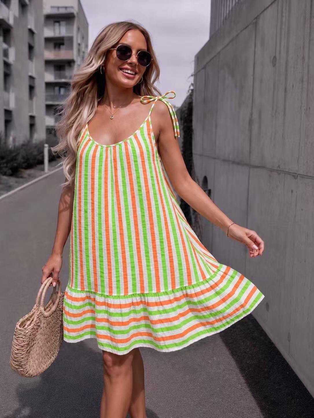 A woman wearing a sleeveless, tiered A-line dress with vertical lime green, orange, and white stripes, walking outdoors on a city street next to a concrete wall.