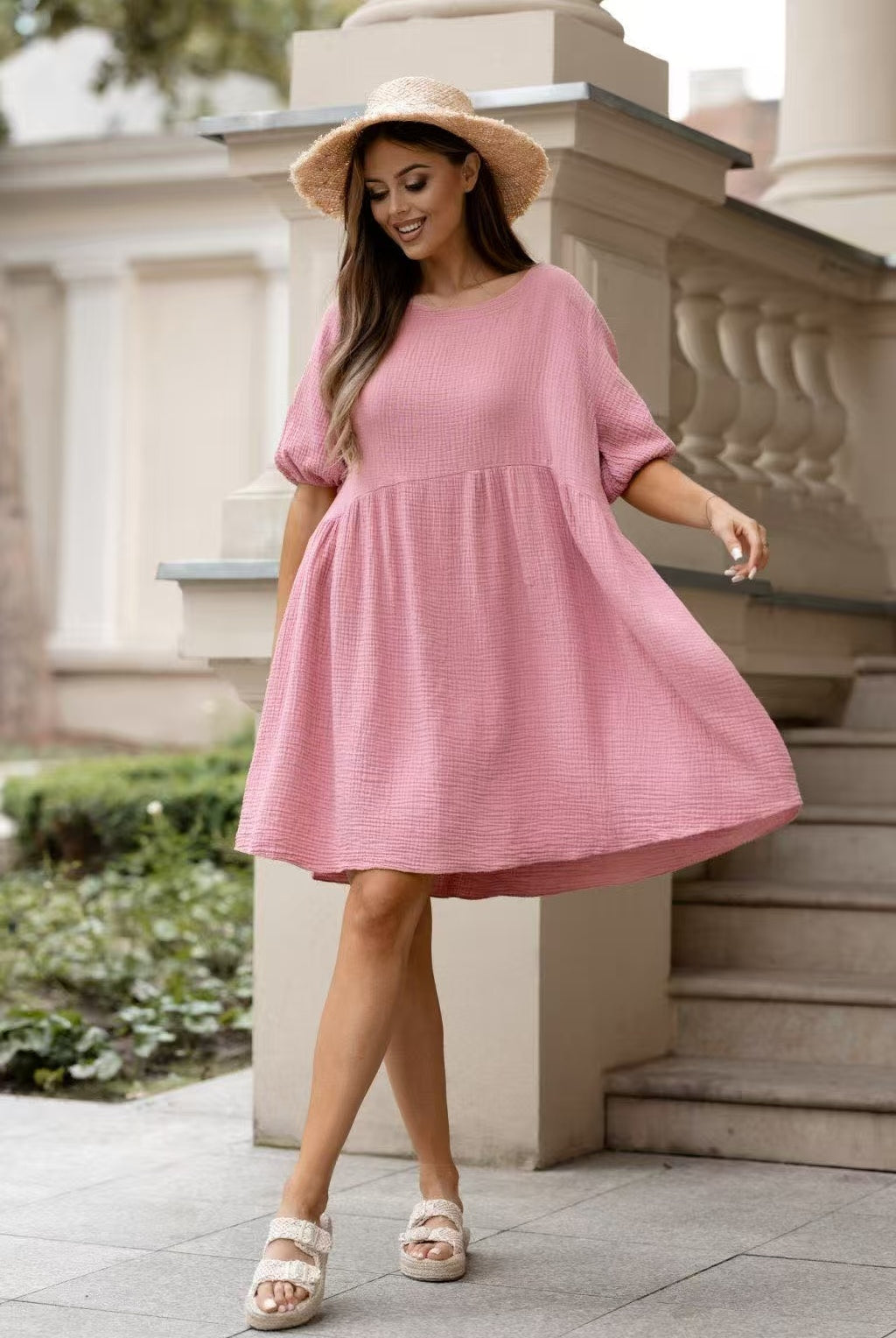 A woman wearing a pink textured short-sleeve dress with a gathered waist, styled with a straw hat and beige platform sandals, standing outdoors in front of a stone balustrade.