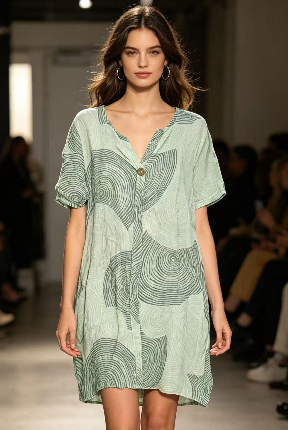 Model wearing a green patterned dress on a runway with an audience.
