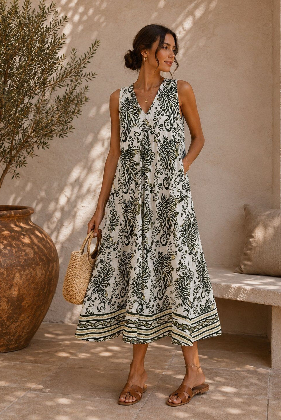 Woman wearing a white dress with olive green patterns standing outdoors near a large vase and bench.
