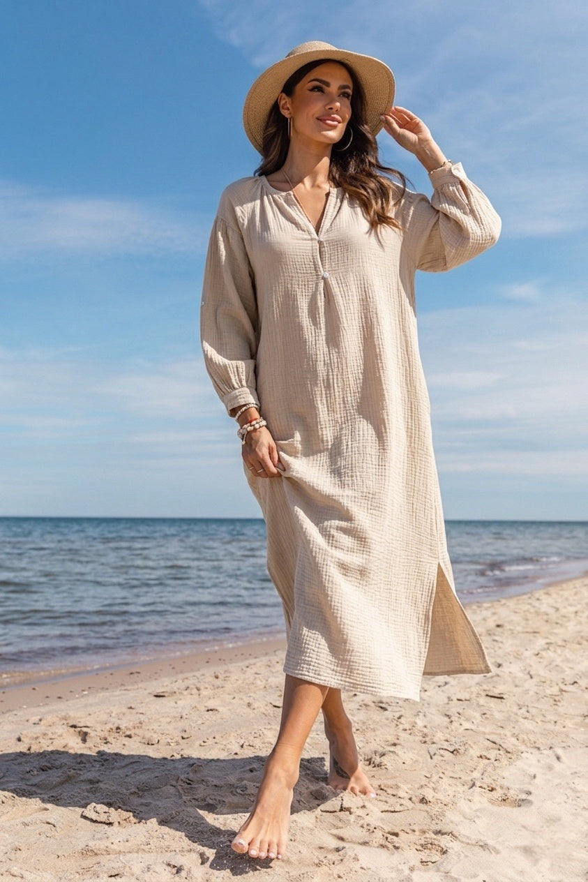 A woman stands on a sandy beach wearing a long-sleeve, midi-length beige textured dress with a V-neck and a wide-brimmed straw hat, with the ocean and blue sky in the background.