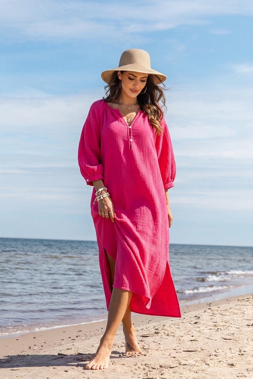 A woman stands on a sandy beach wearing a bright pink maxi cover-up dress with a v-neck and wide sleeves, along with a beige wide-brim hat.