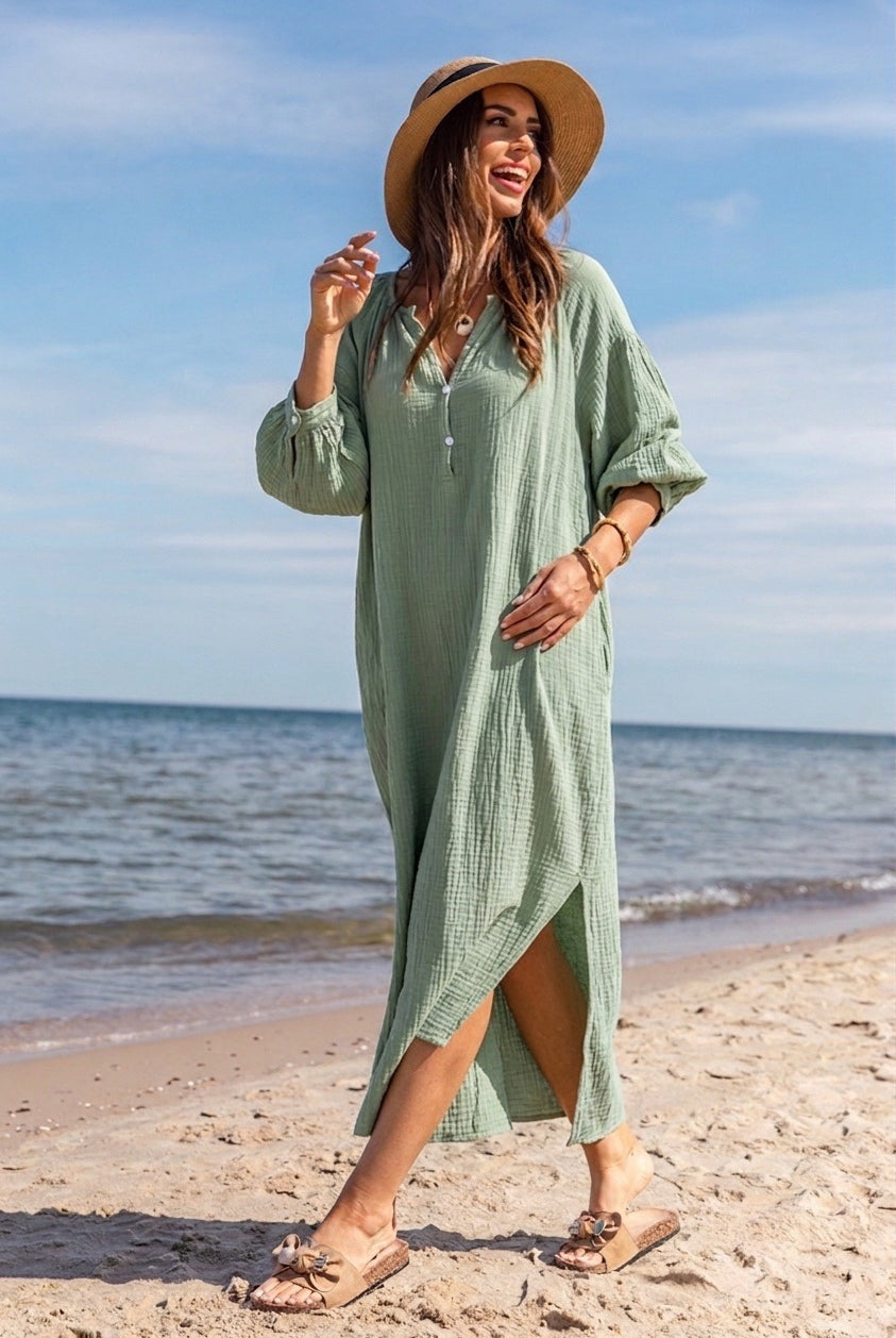A woman wearing a light sage green waffle-knit maxi dress and a wide-brim straw hat walks on a sandy beach with the ocean and blue sky in the background.