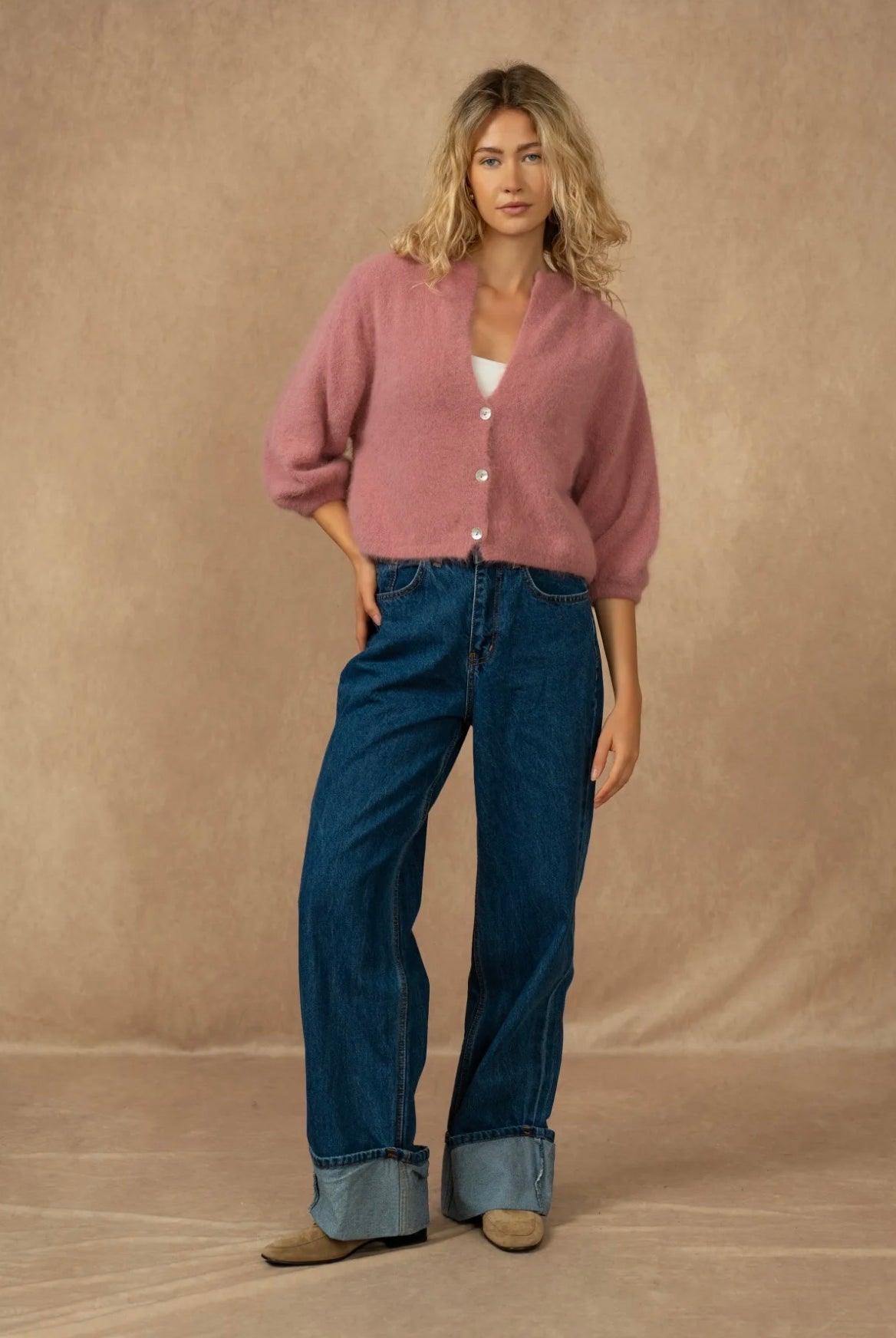 A woman modeling a dusty pink button-up cardigan with three-quarter sleeves, paired with dark blue wide-leg jeans and tan shoes, set against a warm beige studio backdrop.