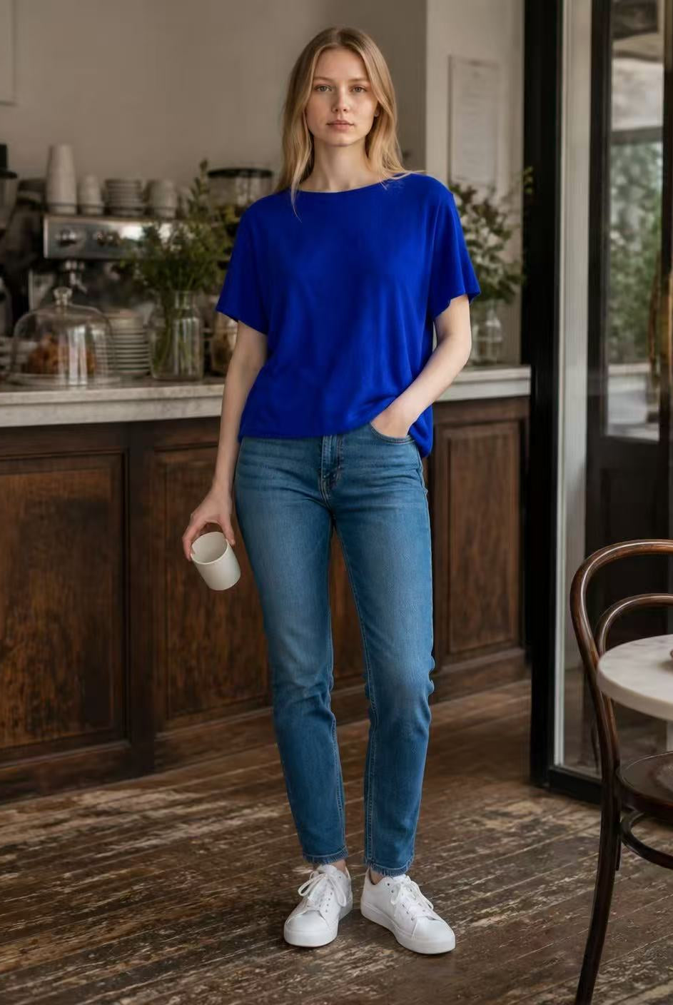 A woman stands in a cafe wearing a vibrant royal blue short-sleeve top, blue jeans, and white sneakers, holding a white mug.