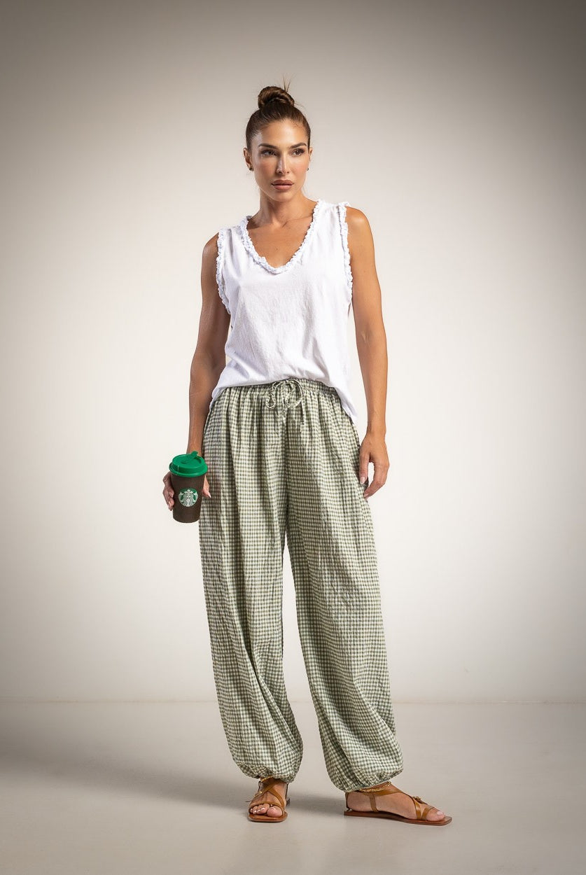 A woman stands against a neutral studio background wearing a white sleeveless top and loose green-and-white checkered pants, holding a dark coffee cup with a green lid.