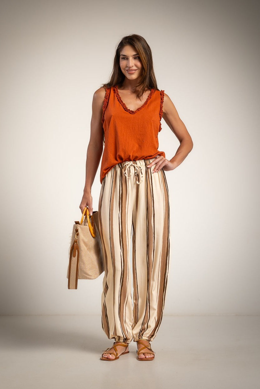A woman stands against a neutral studio backdrop wearing a bright orange sleeveless top and beige-and-brown vertically striped harem-style pants, holding a tan tote bag.