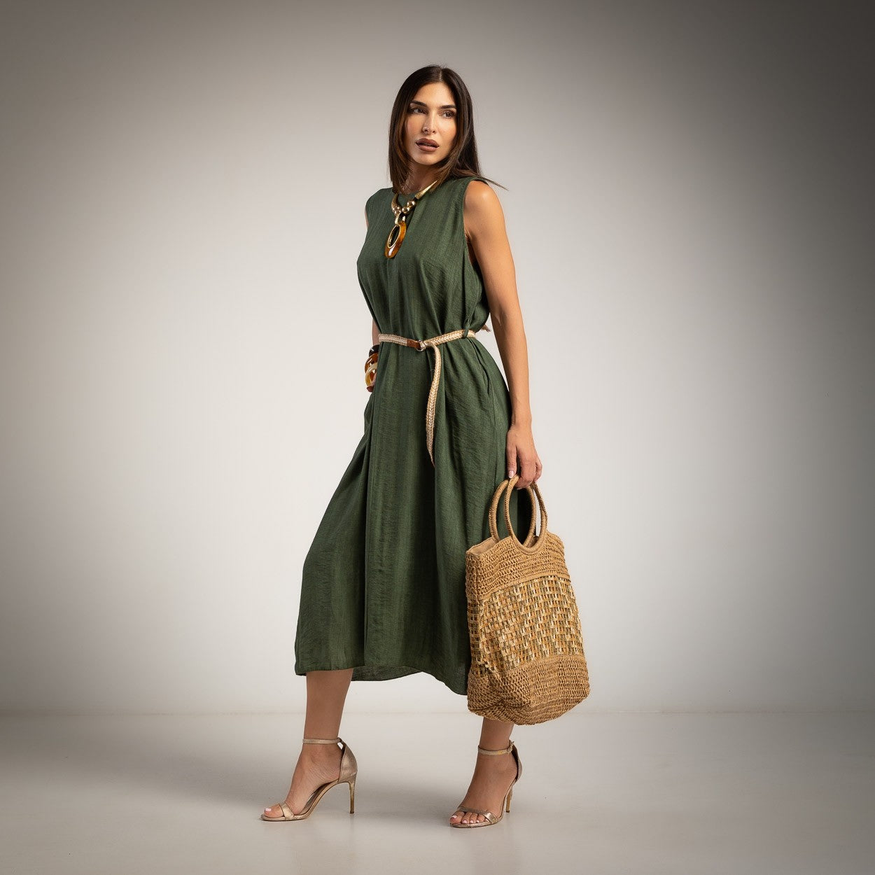 A woman models a sleeveless khaki green midi dress with a beige tie belt, holding a woven straw tote bag