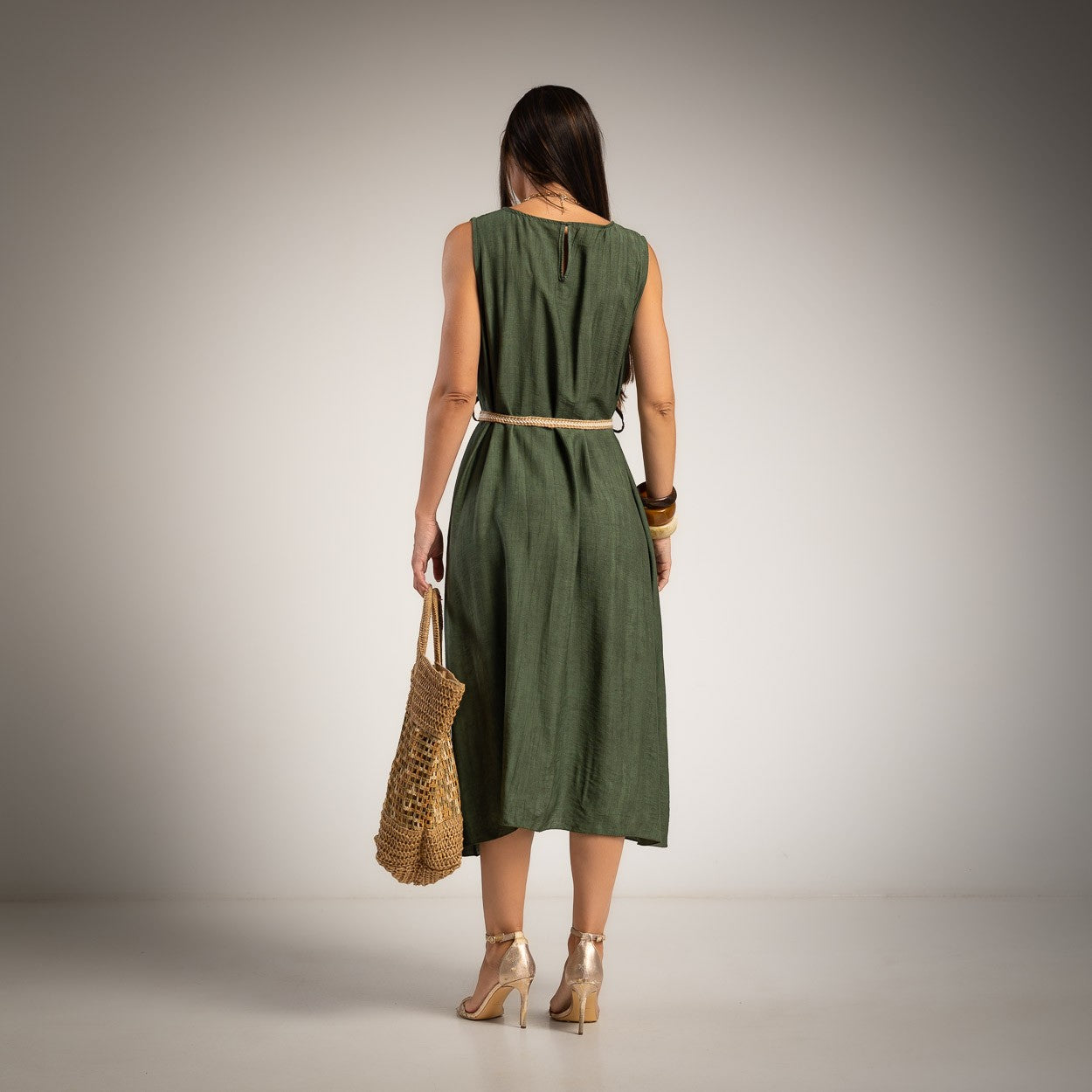 A woman stands with her back to the camera wearing a sleeveless olive green midi dress with a thin beige belt