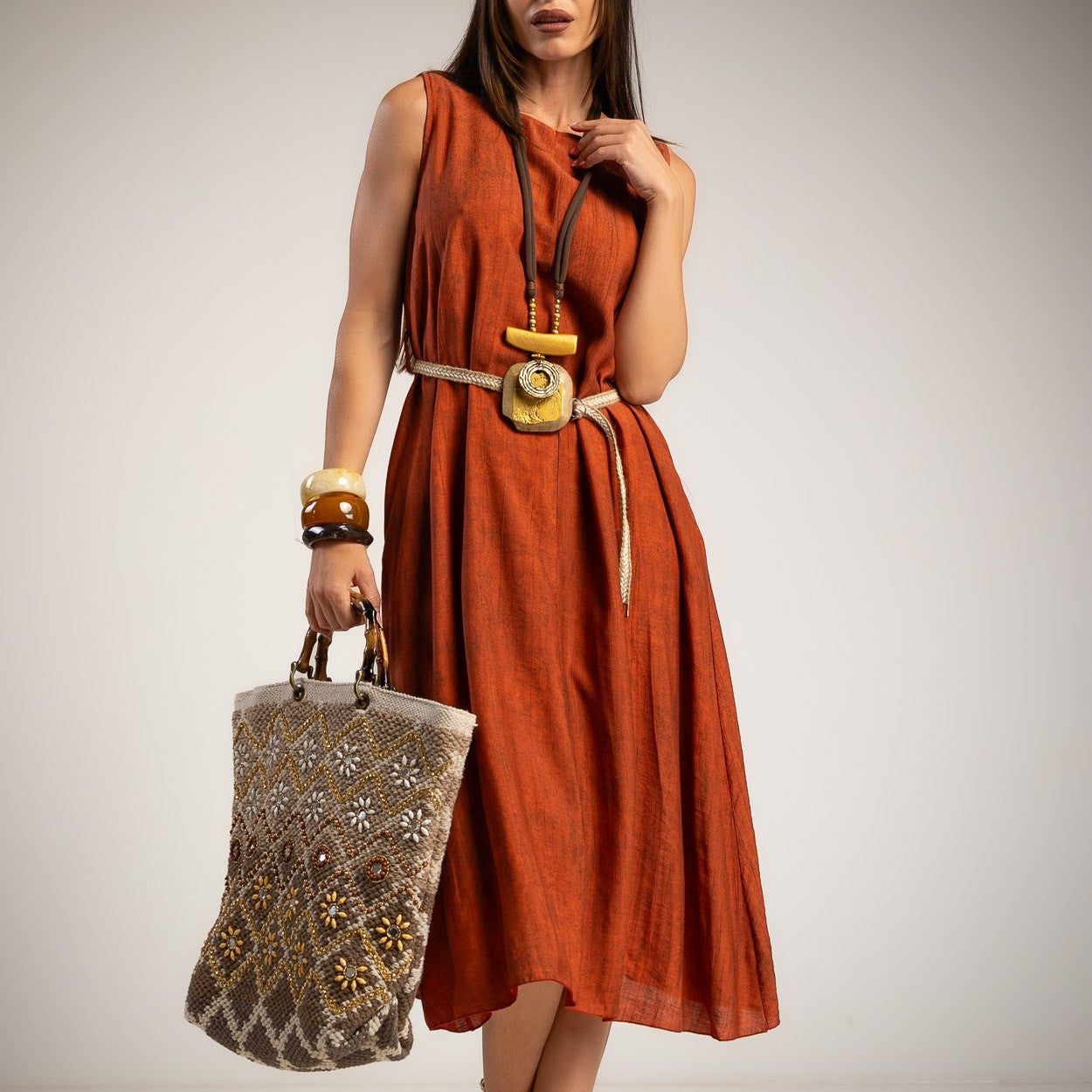 A woman models a sleeveless rust-orange midi dress with a cream belt and a large wooden pendant.