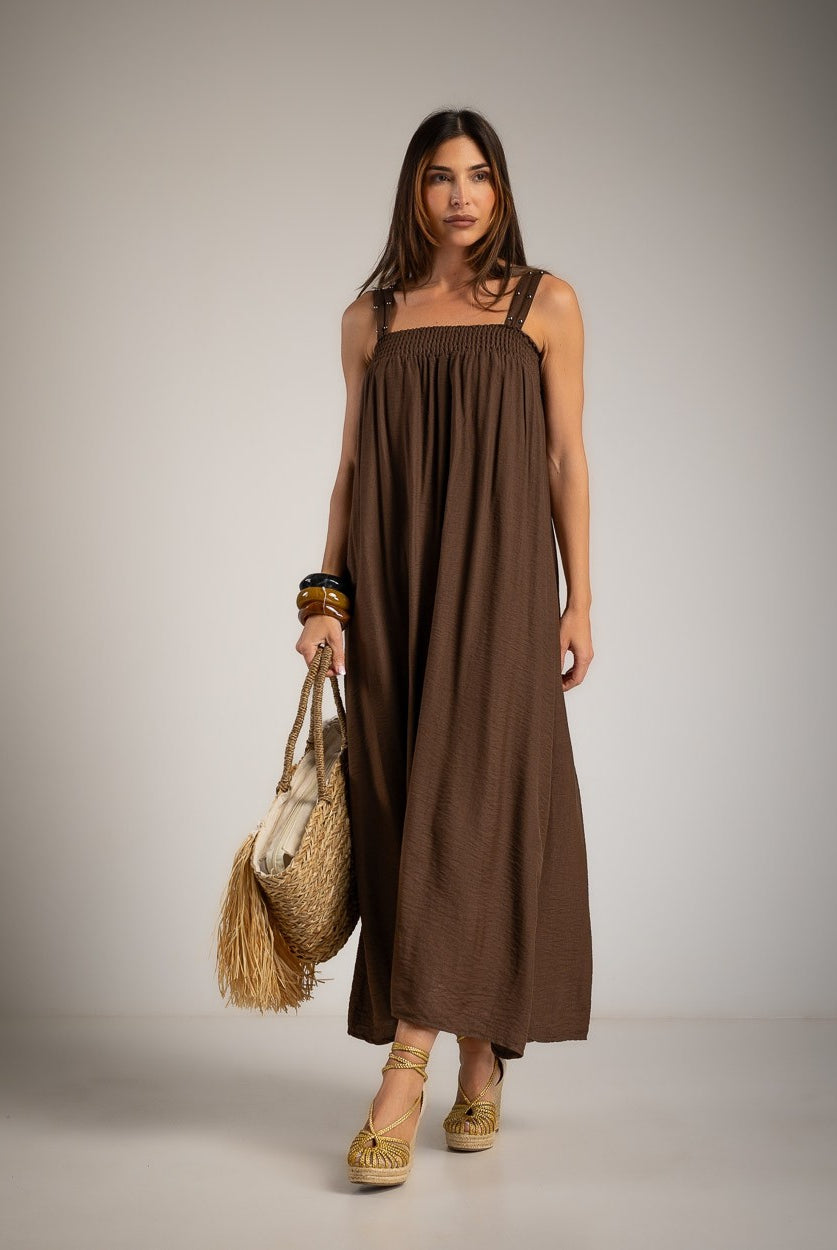 A woman modeling a brown sleeveless dress with a square neckline, holding a straw bag and wearing woven sandals against a neutral studio background.