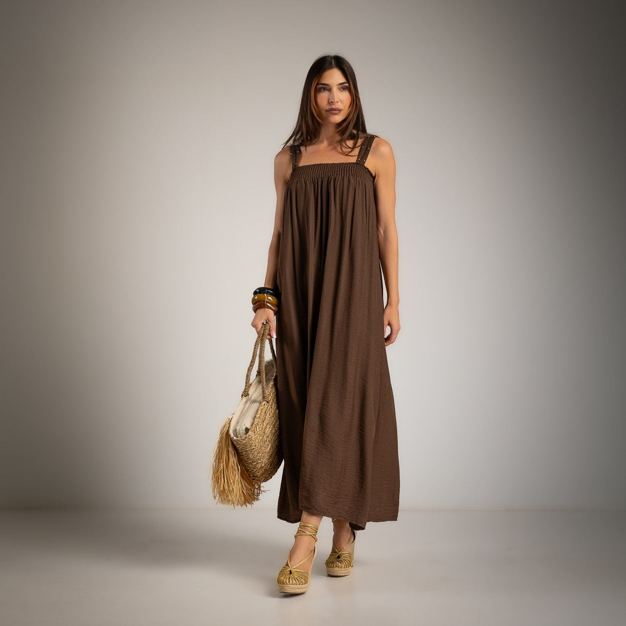 A woman modeling a brown sleeveless dress with a square neckline, holding a straw bag and wearing woven sandals against a neutral studio background.