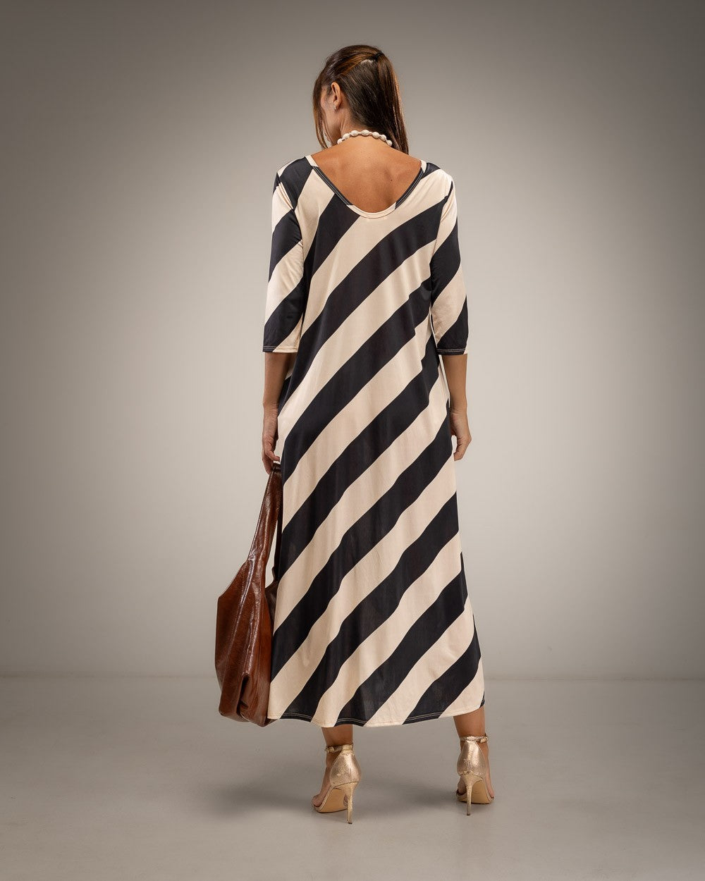 A woman viewed from the back wearing a loose, knee-length dress with bold diagonal black and cream stripes. She is holding a brown leather tote bag