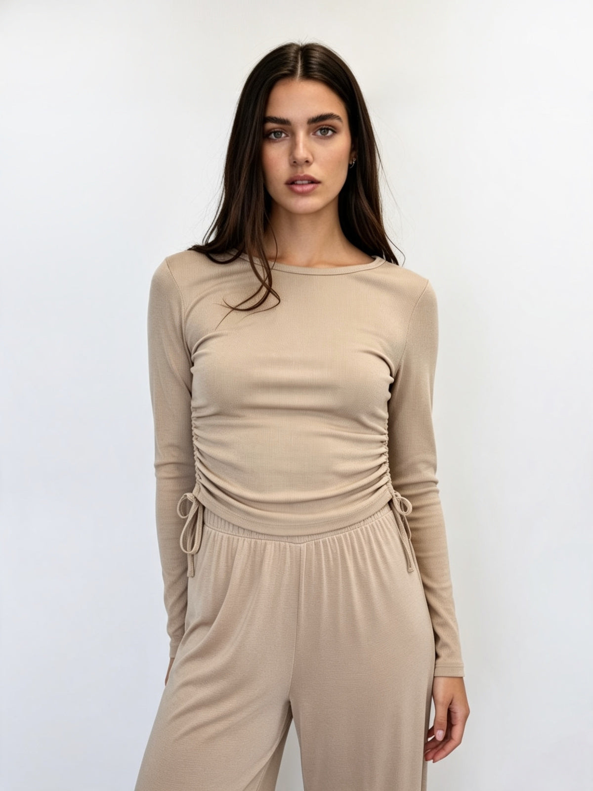 A woman modeling a beige long-sleeve jumpsuit with a ruched bodice and drawstring waist details