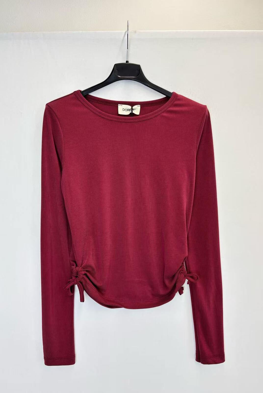 A long-sleeve crewneck top in deep burgundy, hanging on a black hanger against a plain white wall.