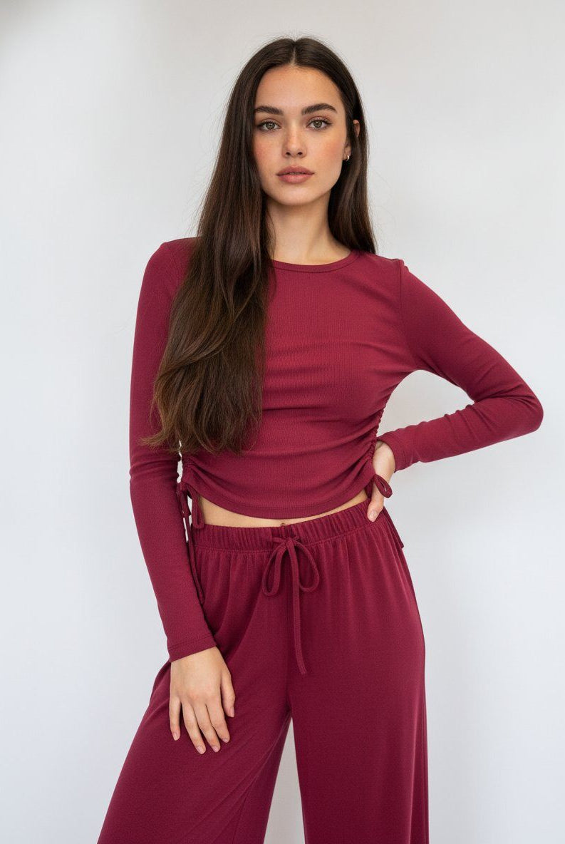 A woman modeling a matching burgundy long-sleeve crop top and drawstring lounge pants against a light gray studio background.