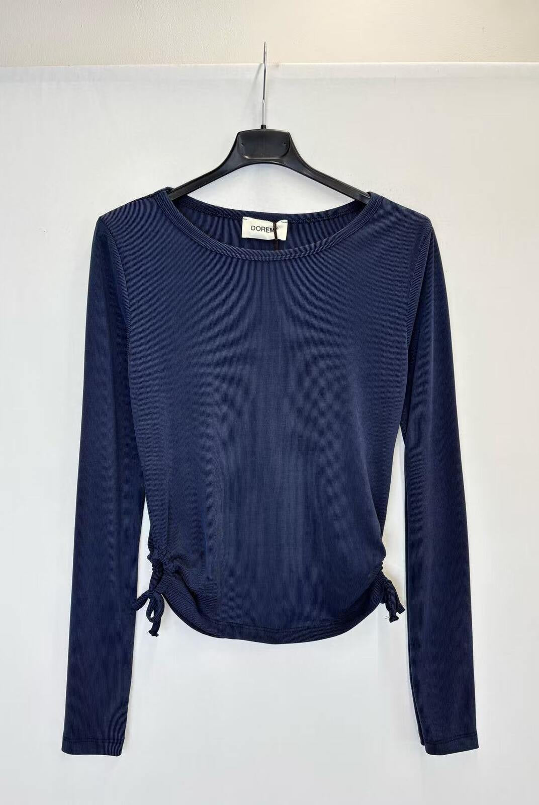 A navy blue long-sleeve knit top hanging on a black hanger against a plain white wall. The top has a crew neckline and side tie details at the hem.