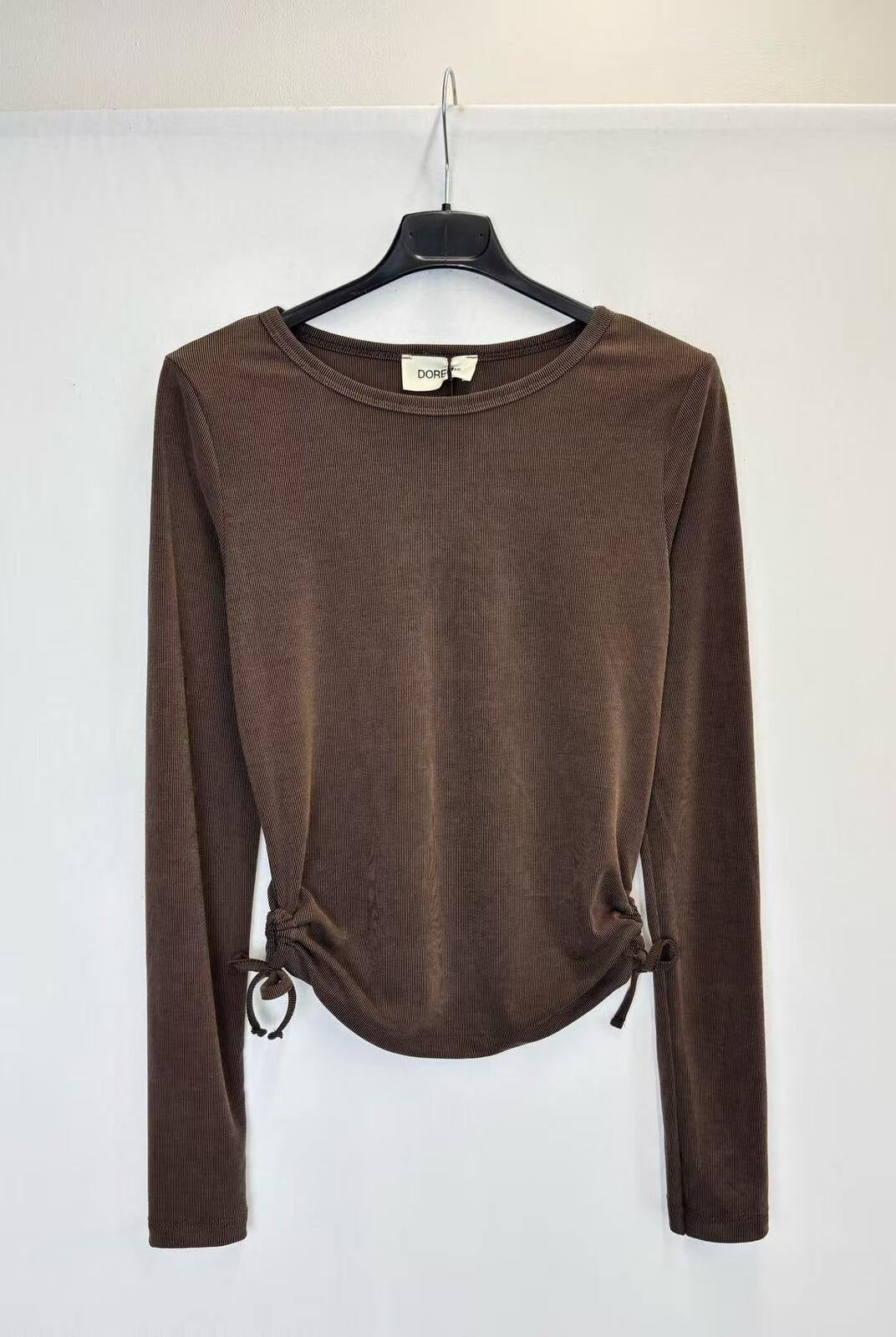 A brown long-sleeve knit top hanging on a black hanger against a plain white wall. The top has a crew neckline and side drawstring details at the hem.
