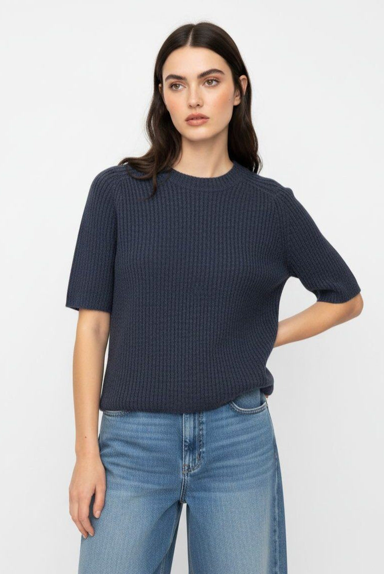 A woman modeling a dark navy blue ribbed short-sleeve knit top with light blue jeans, standing against a plain white studio background.
