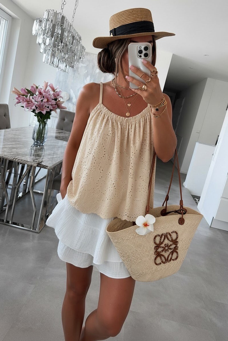 A woman takes a mirror selfie in a modern, bright room, wearing a beige eyelet camisole top, a white ruffled skirt, a straw hat, and carrying a beige woven tote bag with a white flower accent.