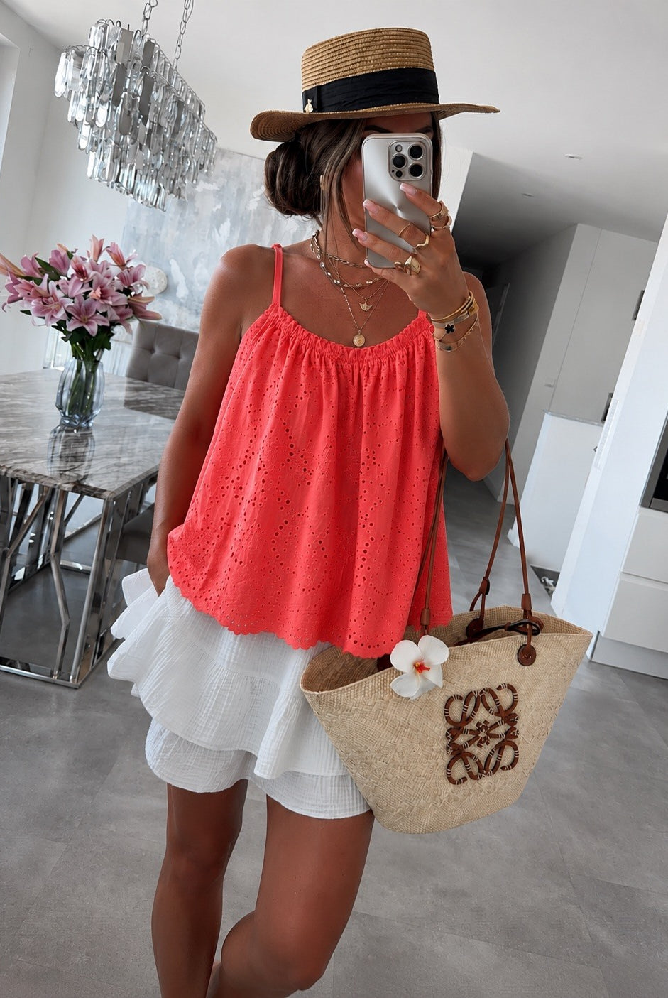 A woman takes a mirror selfie in a modern dining room, wearing a bright coral pink eyelet spaghetti-strap top, a white ruffled mini skirt, a straw hat with a black band, and holding a beige woven straw tote bag with a white flower accent.