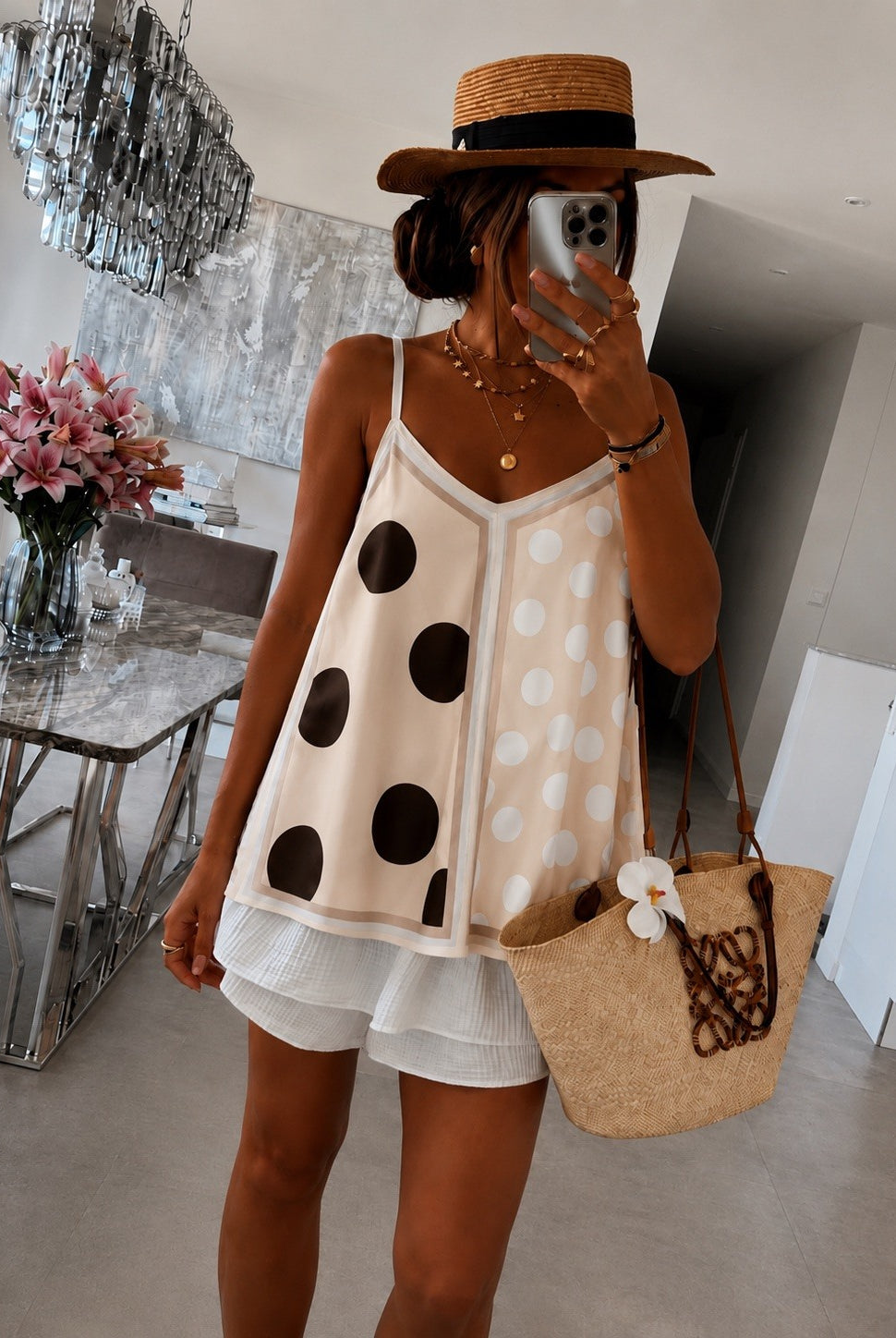 A woman takes a mirror selfie wearing a beige camisole top with large black and white polka dots, white ruffled shorts, a straw hat, and carrying a woven straw tote bag in a modern indoor setting.