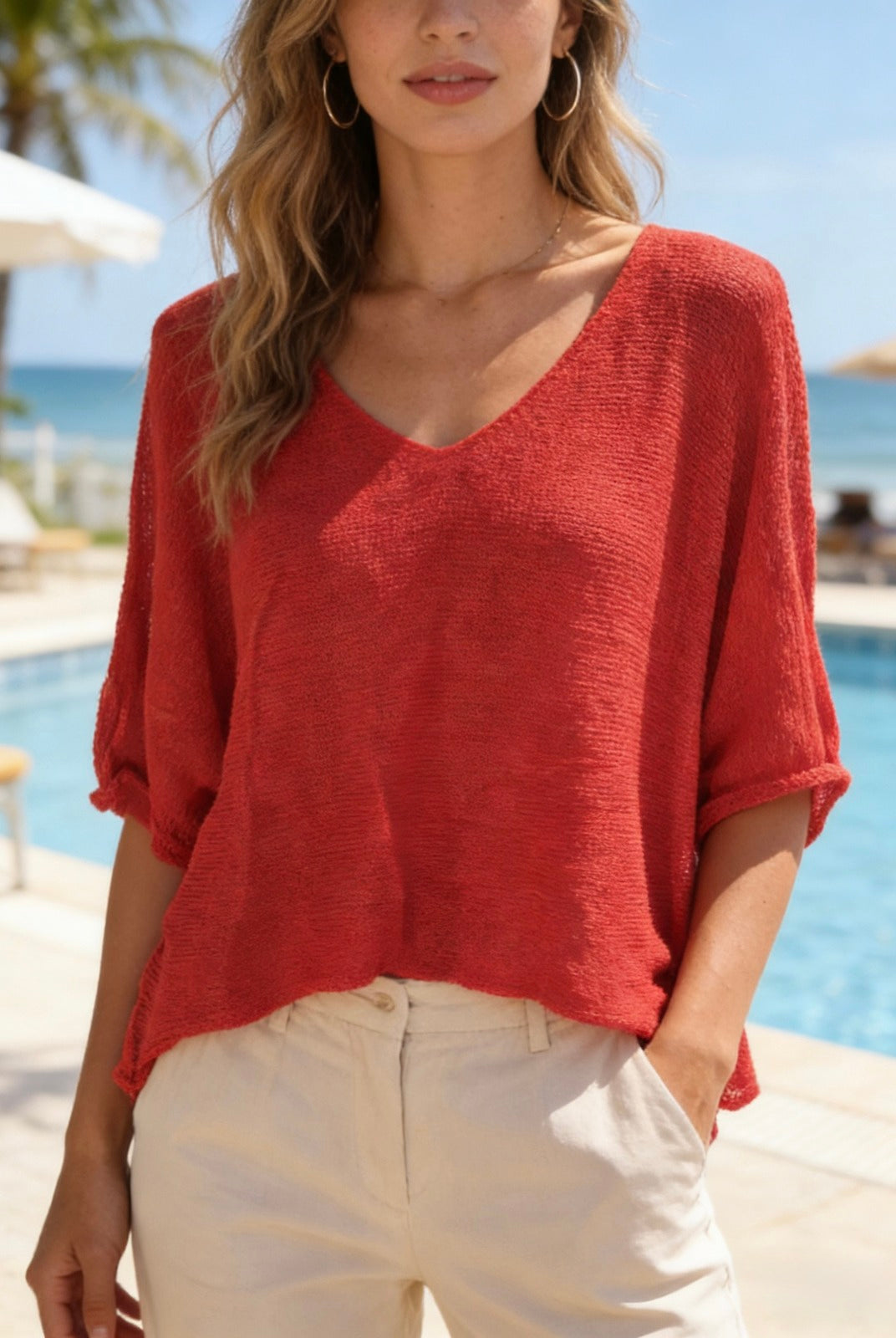 Woman wearing a red top and beige pants by a pool with palm trees in the background