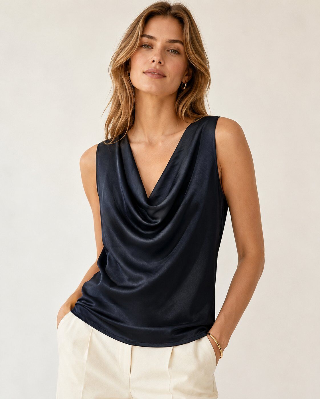 A woman wearing a sleeveless navy blue cowl-neck top with a satin-like sheen, paired with cream-colored pants, against a plain light beige background.