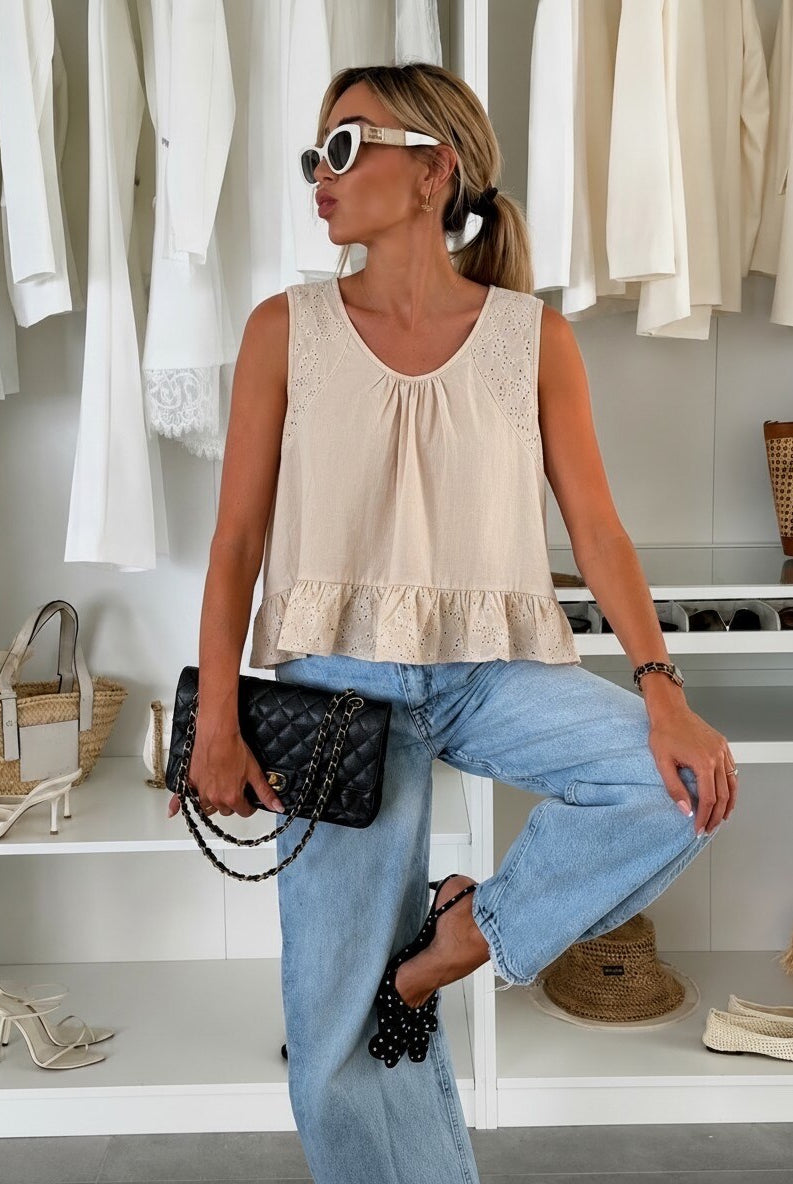 A woman models a sleeveless beige top with a ruffled hem, paired with light blue jeans and a black quilted clutch bag, set against a background of white clothing racks.