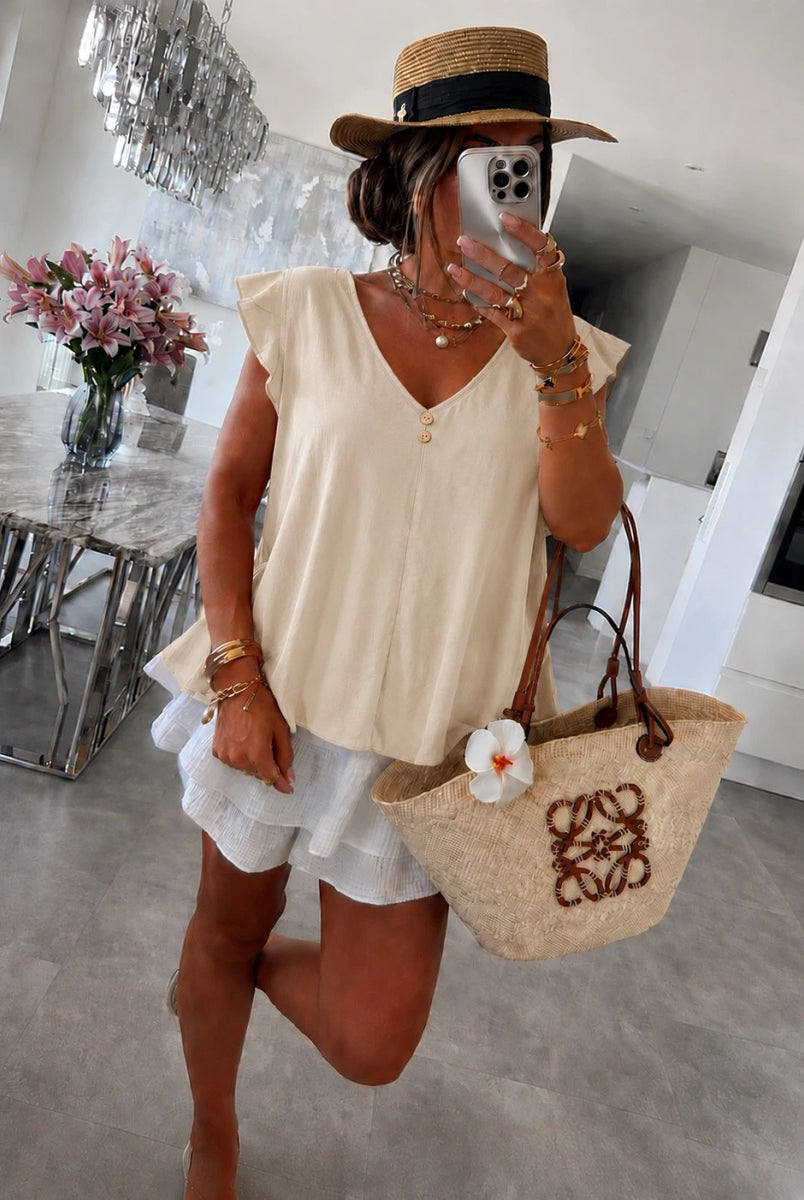A woman wearing a cream sleeveless V-neck top, white ruffled shorts, a straw hat with a black band, and carrying a beige woven tote bag