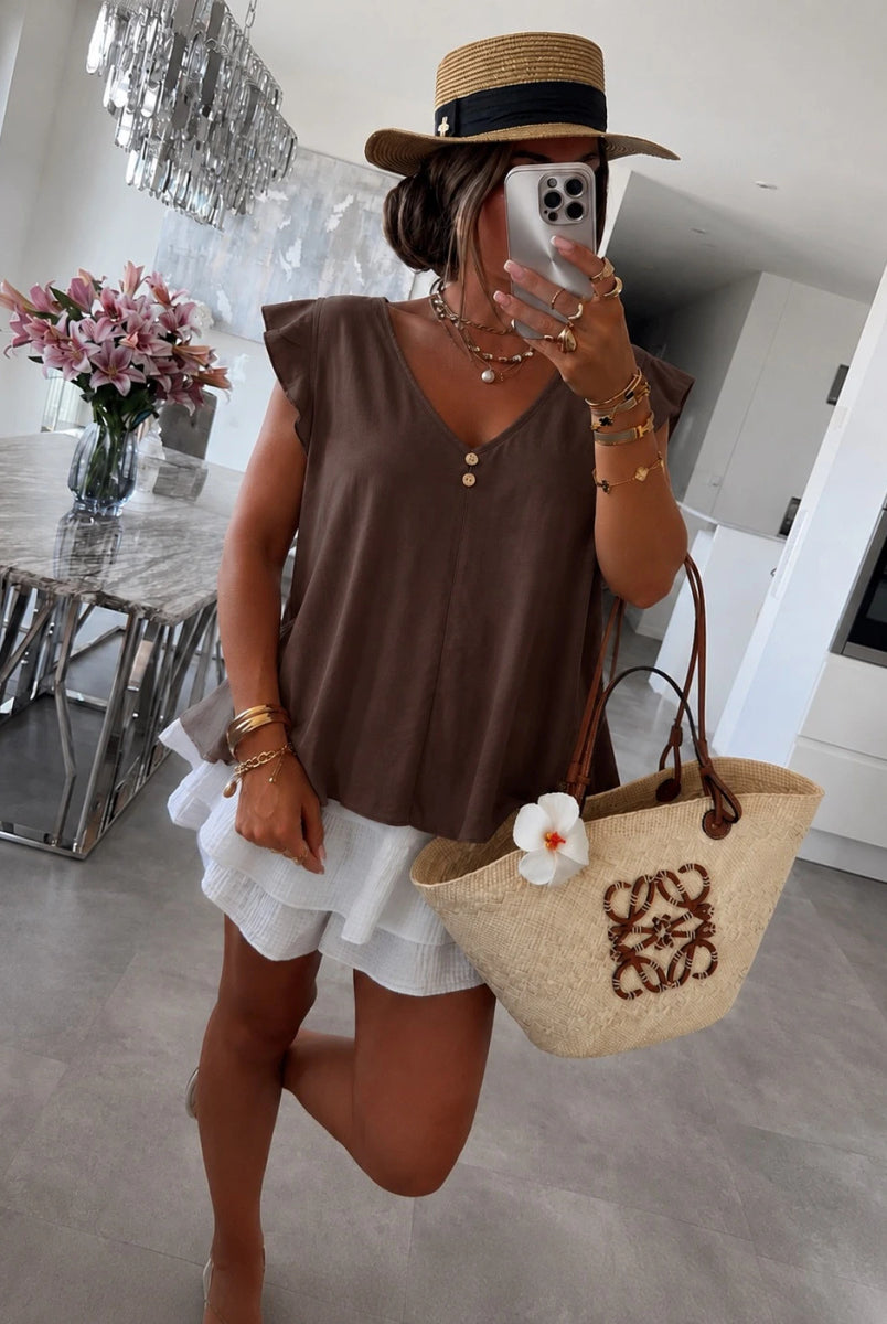 A woman wearing a brown sleeveless V-neck top, white ruffled shorts, a straw hat, and carrying a beige woven tote bag