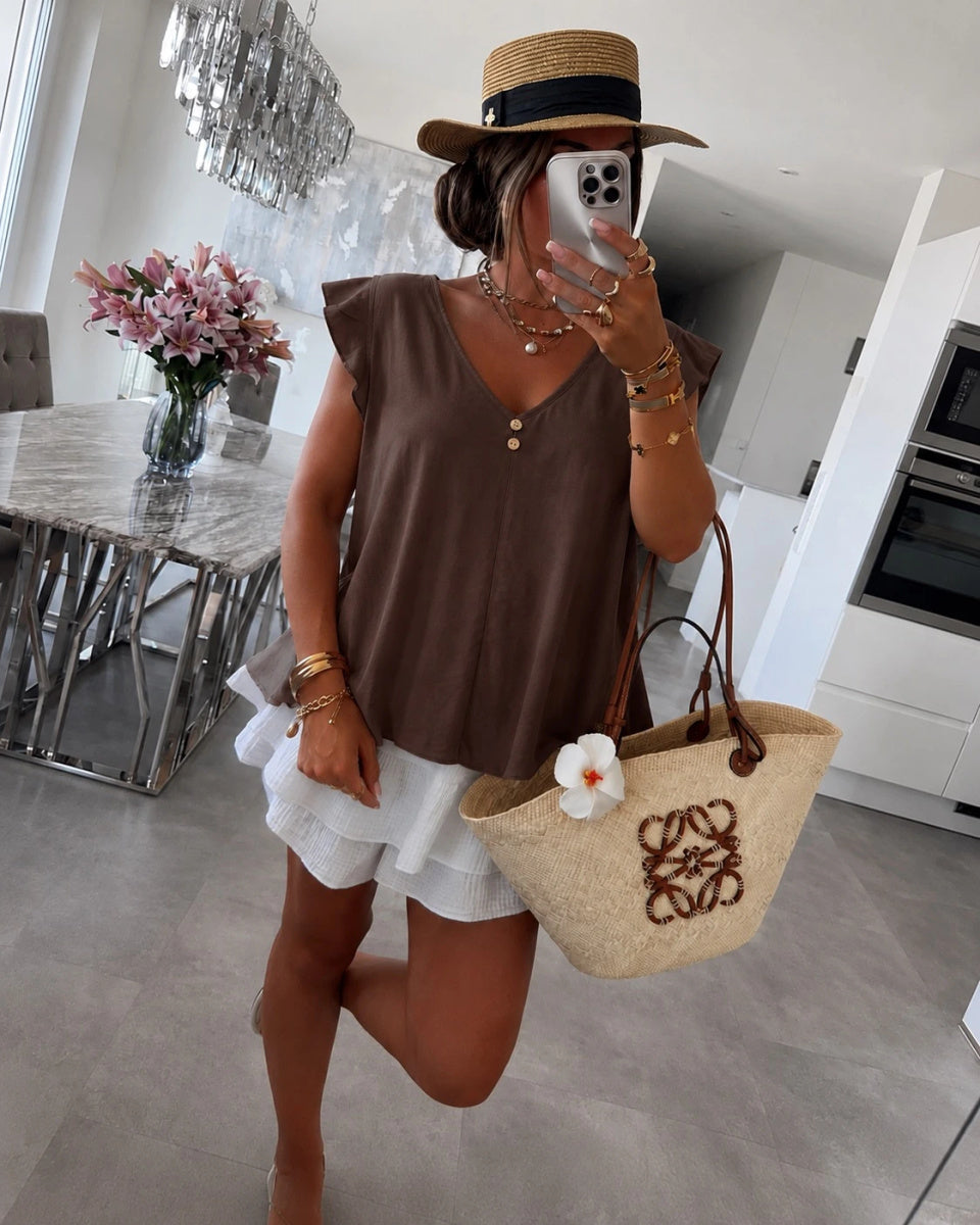 A woman wearing a brown sleeveless V-neck top, white ruffled shorts, a straw hat, and carrying a beige woven tote bag