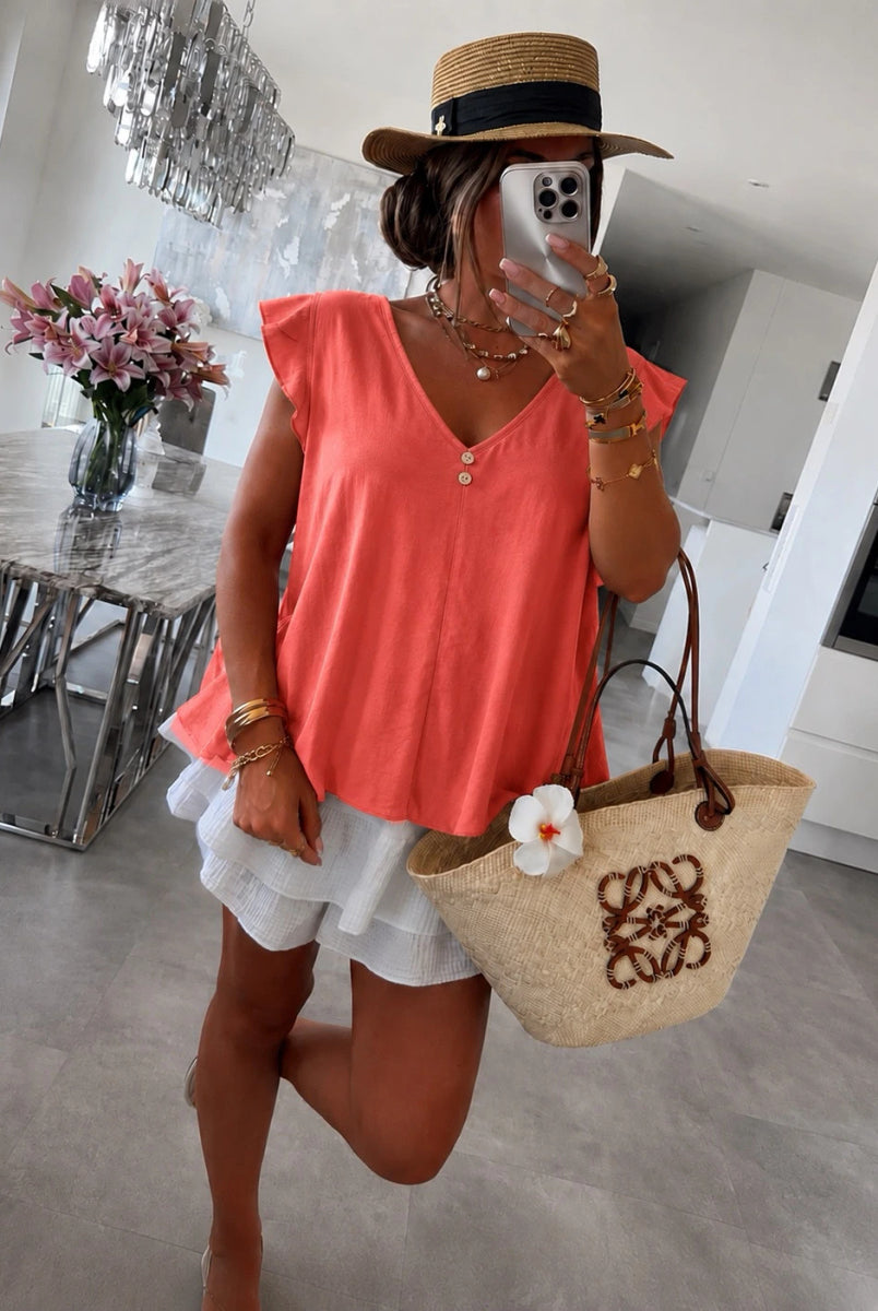 A woman wearing a coral sleeveless top, white ruffled shorts, a straw hat, and carrying a beige woven tote bag